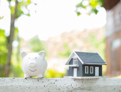 Tirelire blanche en forme de cochon et maquette de masion dans une foret repr&eacute;sentant les assurance de pret immobilier