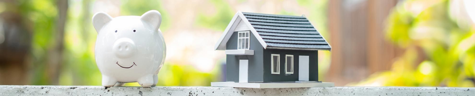 Maquette d'une maison a cot&eacute; d'une tirelire en forme de cochon blanc sur fonc foret Assurance Pr&ecirc;t immobilier